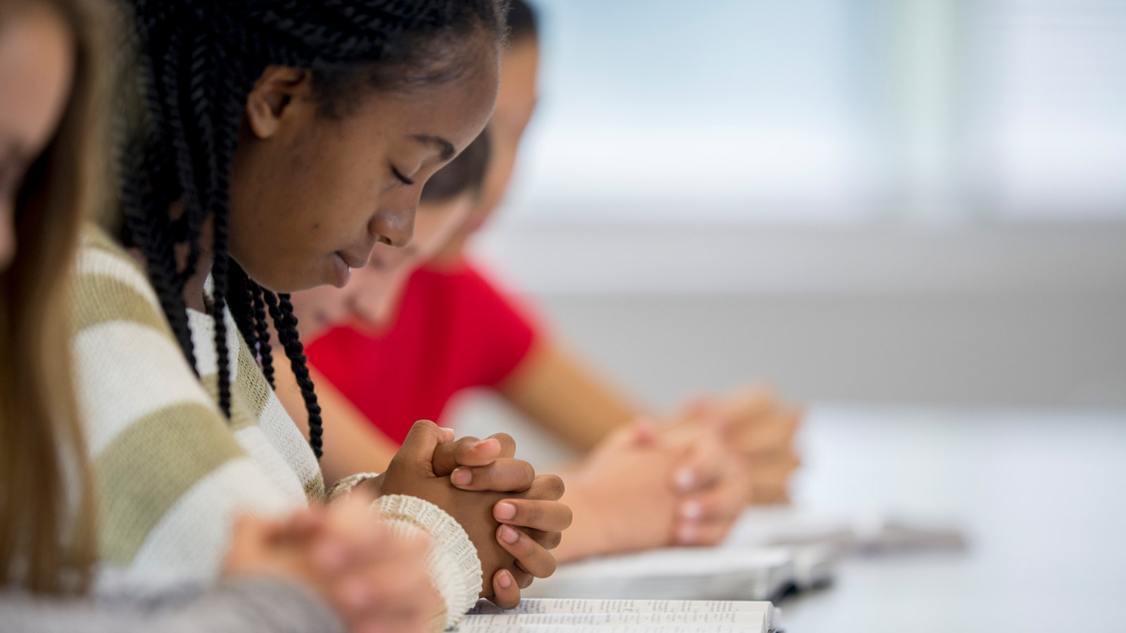 Students with hands crossed on Bibles and heads bowed in prayer.